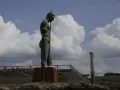 Brązowy posąg mężczyzny z włócznią na tle nieba z chmurami i ruinami Pompejów.