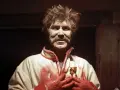 Mężczyzna z twarzą pokrytą białą substancją, w białej kurtce z czerwonymi zdobieniami i medalionem. Jego dłonie są czerwone, jakby od krwi. Może to symbolizować trudne wesele, gdzie kaduceusz symbolizuje pokój, ale tu jest złamany.