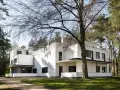 Biała, modernistyczna willa, przykład bauhaus architektury, otoczona drzewami.