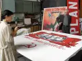 Kobieta maluje na wielkim płótnie z podobiznami przywódców i sierpem i młotem. W tle mężczyzna stoi obok plakatu z napisem "PLAN". Powidok historii.