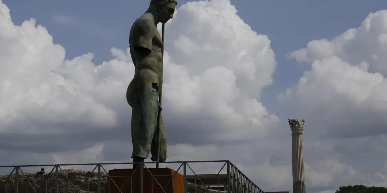 Brązowy posąg mężczyzny z włócznią na tle nieba z chmurami i ruinami Pompejów.