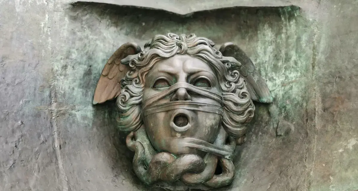 Brązowa rzeźba głowy z opaską na oczach i ustach, ze skrzydłami i wężami.