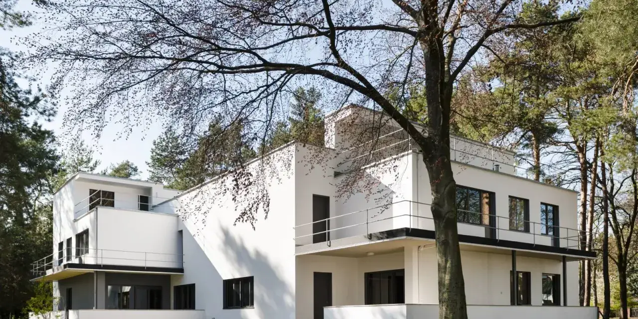 Biała, modernistyczna willa, przykład bauhaus architektury, otoczona drzewami.