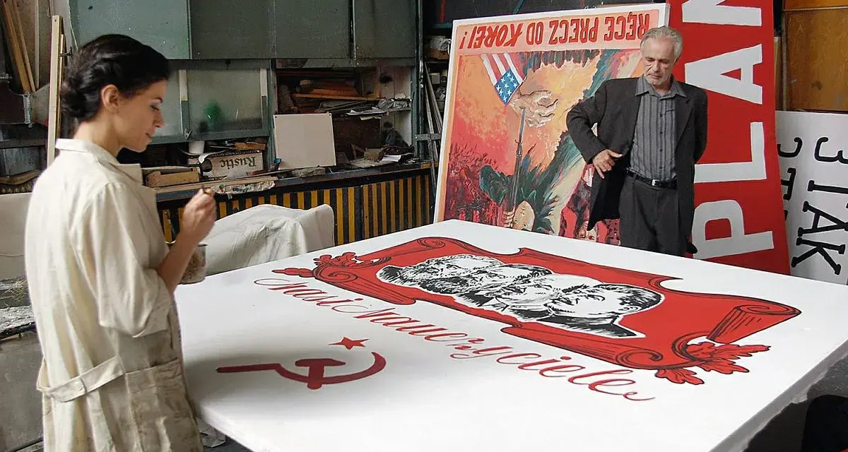 Kobieta maluje na wielkim płótnie z podobiznami przywódców i sierpem i młotem. W tle mężczyzna stoi obok plakatu z napisem "PLAN". Powidok historii.