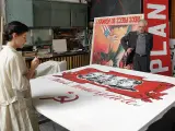 Kobieta maluje na wielkim płótnie z podobiznami przywódców i sierpem i młotem. W tle mężczyzna stoi obok plakatu z napisem "PLAN". Powidok historii.