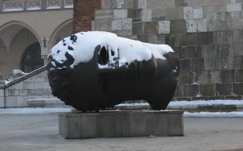 Duża, ciemna rzeźba głowy pokryta śniegiem, na tle kamiennej ściany i arkadowego budynku.