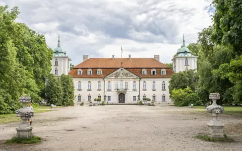 Pałac w Nieborowie, biała rezydencja z czerwonym dachem, otoczona zielenią. Dwie wieżyczki z zielonymi kopułami po bokach.