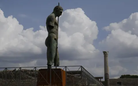 Brązowy posąg mężczyzny z włócznią na tle nieba z chmurami i ruinami Pompejów.