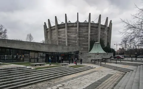 Monumentalny budynek w stylu brutalizm z betonowymi żebrami i szklaną fasadą. Schody prowadzą do wejścia, a wokół zielone trawniki.