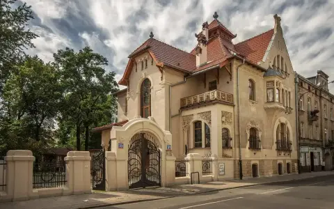 Elegancka willa w stylu art nouveau z bogatymi zdobieniami, kutą bramą i ozdobnym balkonem.