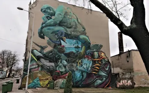 Co to jest mural? Ogromna, surrealistyczna grafika na ścianie budynku, przedstawiająca myślącą postać, krajobraz morski, jedzenie i przedmioty codziennego użytku.