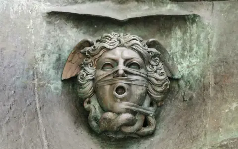 Brązowa rzeźba głowy z opaską na oczach i ustach, ze skrzydłami i wężami.