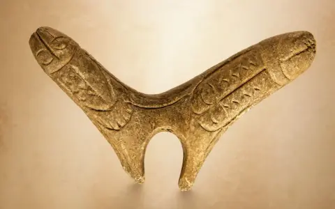 Starożytny, rzeźbiony w drewnie fallus, przypominający stylizowanego ptaka lub zwierzę, z wyrytymi detalami.