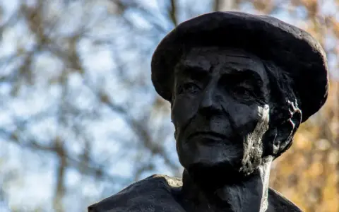 Portret rzeźbiarski w ciemnym brązie, ukazujący twarz mężczyzny w beretce. Widać głębokie zmarszczki i wyraziste rysy, oddające estetykę dzieł Kenara.
