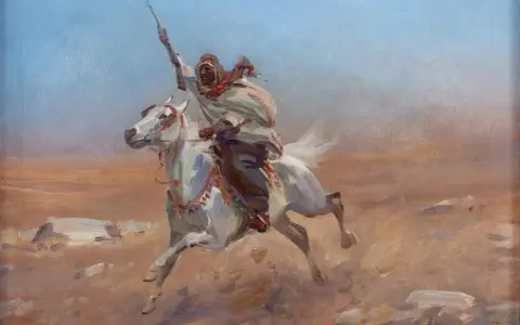 Biały koń galopuje przez pustynię, dosiada go wojownik w tradycyjnym stroju, z uniesioną włócznią. Obraz w stylu orientalizm.