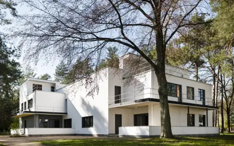 Biała, modernistyczna willa, przykład bauhaus architektury, otoczona drzewami.