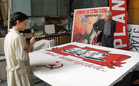 Kobieta maluje na wielkim płótnie z podobiznami przywódców i sierpem i młotem. W tle mężczyzna stoi obok plakatu z napisem "PLAN". Powidok historii.