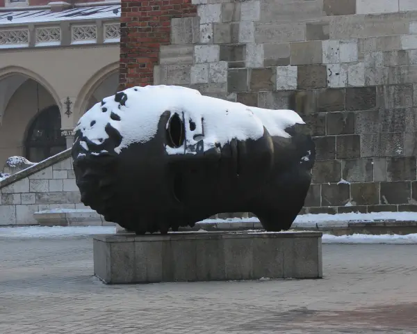 Duża, ciemna rzeźba głowy pokryta śniegiem, na tle kamiennej ściany i arkadowego budynku.