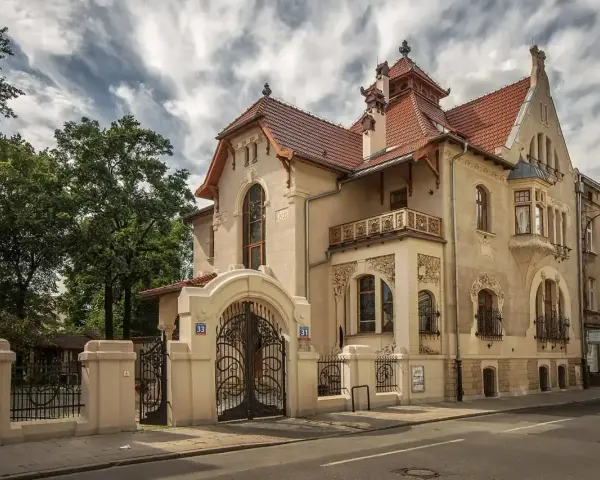 Elegancka willa w stylu art nouveau z bogatymi zdobieniami, kutą bramą i ozdobnym balkonem.