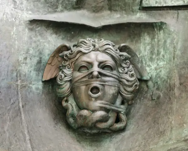 Brązowa rzeźba głowy z opaską na oczach i ustach, ze skrzydłami i wężami.
