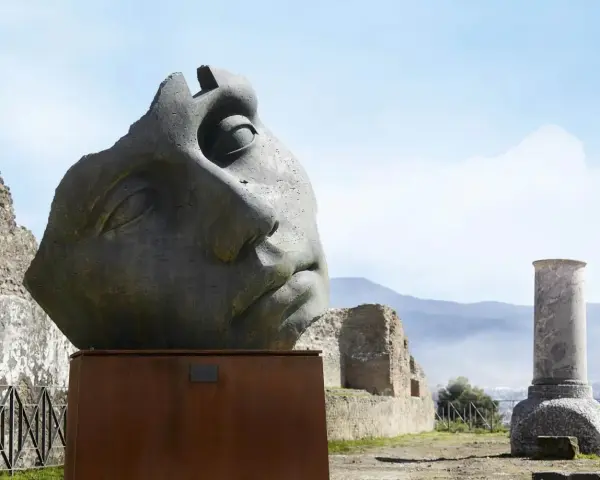 Kamienna rzeźba głowy z pęknięciami na tle ruin i gór.
