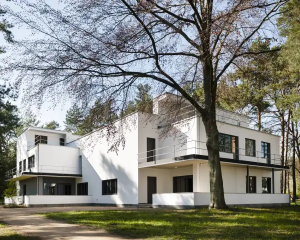 Biała, modernistyczna willa, przykład bauhaus architektury, otoczona drzewami.