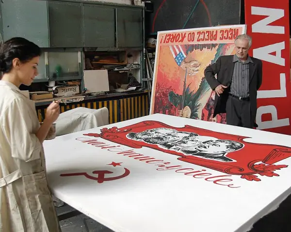 Kobieta maluje na wielkim płótnie z podobiznami przywódców i sierpem i młotem. W tle mężczyzna stoi obok plakatu z napisem "PLAN". Powidok historii.