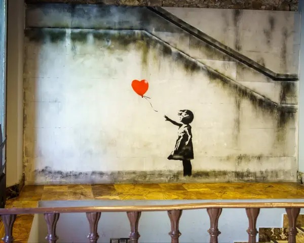 Banksy dziewczynka z balonikiem, unoszącym się na tle schodów i zniszczonej ściany.