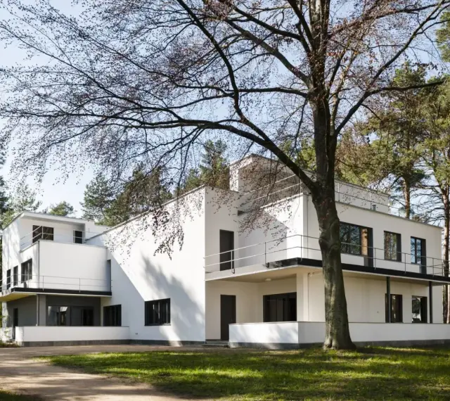 Biała, modernistyczna willa, przykład bauhaus architektury, otoczona drzewami.