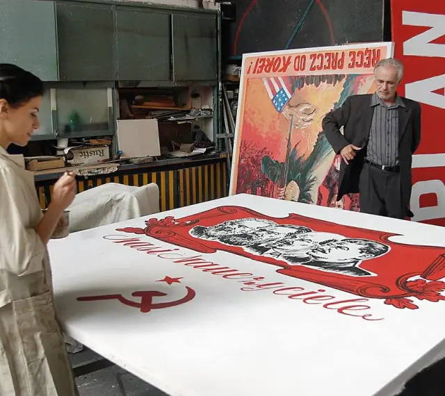 Kobieta maluje na wielkim płótnie z podobiznami przywódców i sierpem i młotem. W tle mężczyzna stoi obok plakatu z napisem "PLAN". Powidok historii.