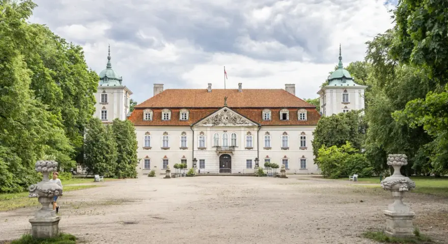 Pałac w Nieborowie, biała rezydencja z czerwonym dachem, otoczona zielenią. Dwie wieżyczki z zielonymi kopułami po bokach.