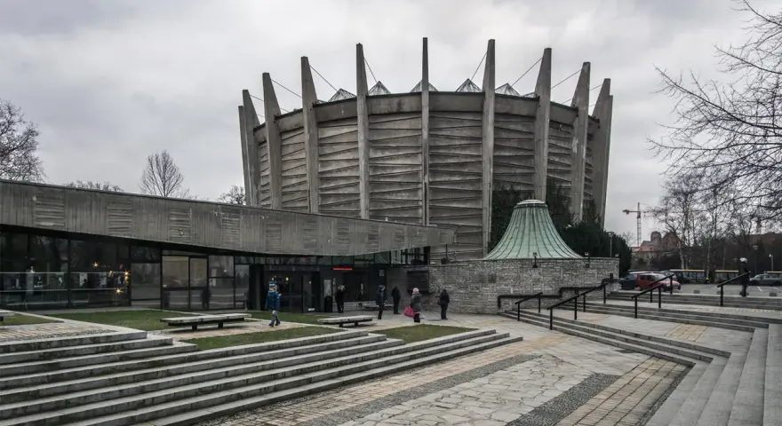 Monumentalny budynek w stylu brutalizm z betonowymi żebrami i szklaną fasadą. Schody prowadzą do wejścia, a wokół zielone trawniki.
