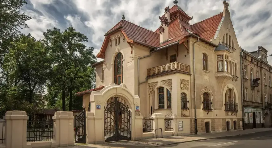 Elegancka willa w stylu art nouveau z bogatymi zdobieniami, kutą bramą i ozdobnym balkonem.