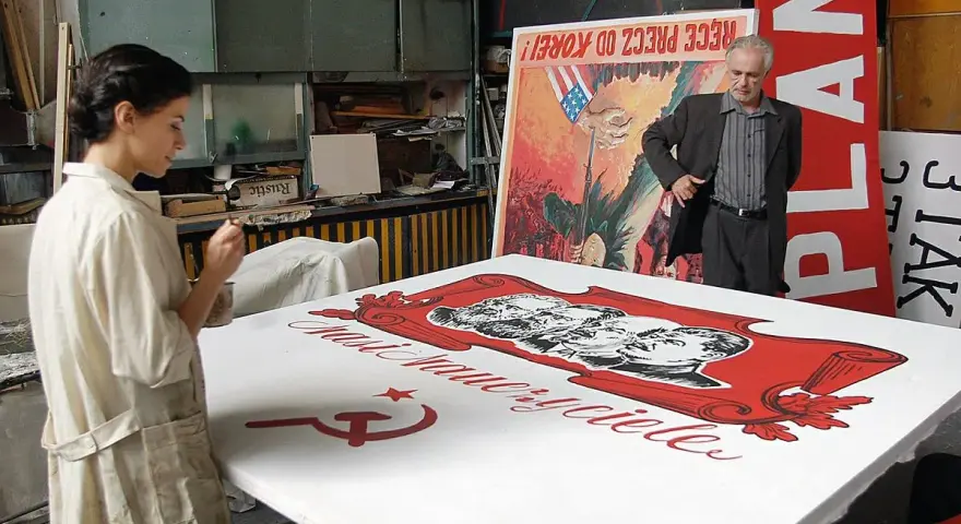 Kobieta maluje na wielkim płótnie z podobiznami przywódców i sierpem i młotem. W tle mężczyzna stoi obok plakatu z napisem "PLAN". Powidok historii.