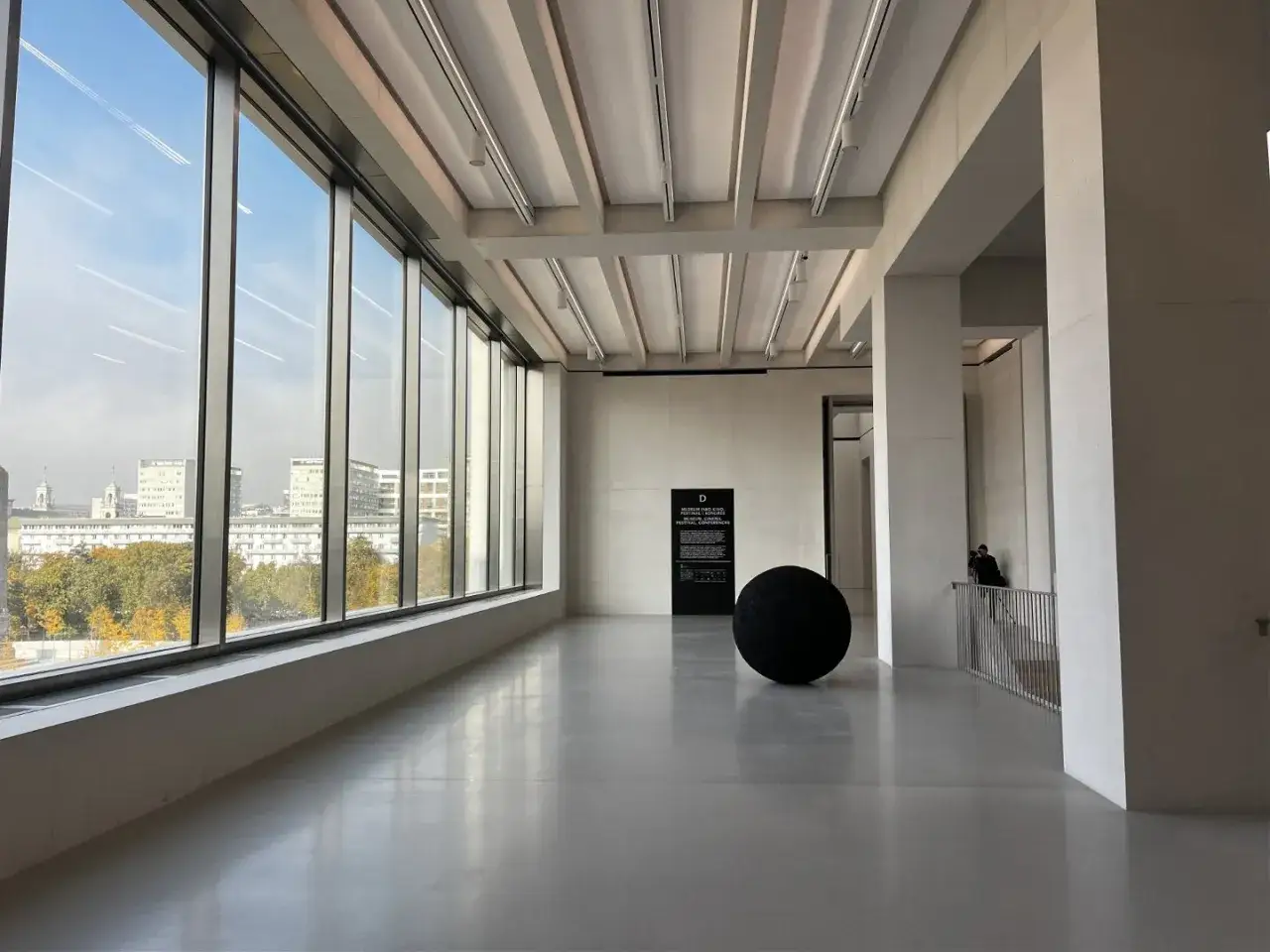 Wnętrze muzeum sztuki nowoczesnej w Warszawie. Duża czarna kula jako instalacja, za nią tablica informacyjna. Okna wychodzą na miasto.