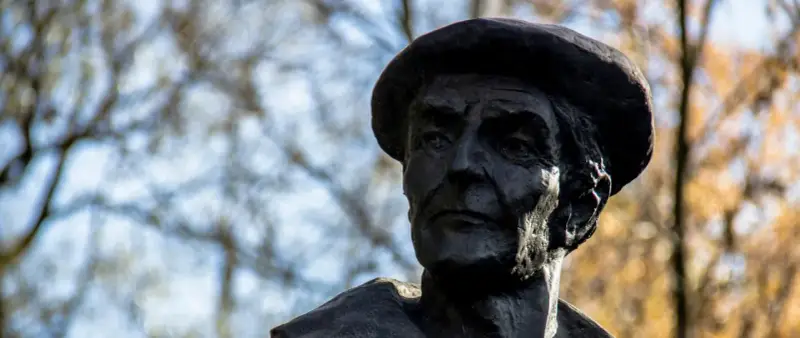 Portret rzeźbiarski w ciemnym brązie, ukazujący twarz mężczyzny w beretce. Widać głębokie zmarszczki i wyraziste rysy, oddające estetykę dzieł Kenara.