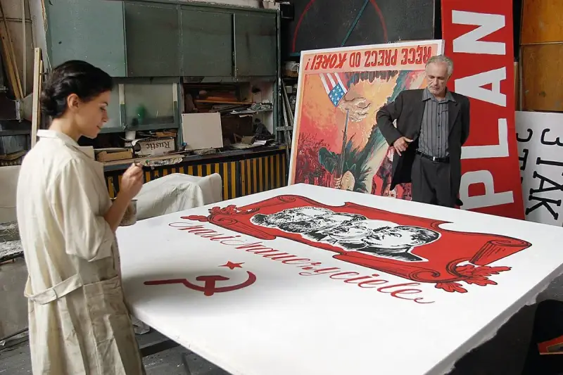 Kobieta maluje na wielkim płótnie z podobiznami przywódców i sierpem i młotem. W tle mężczyzna stoi obok plakatu z napisem "PLAN". Powidok historii.